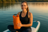 headshot image: Woman on paddleboard holding the bag with a smile, calm lake in the background.